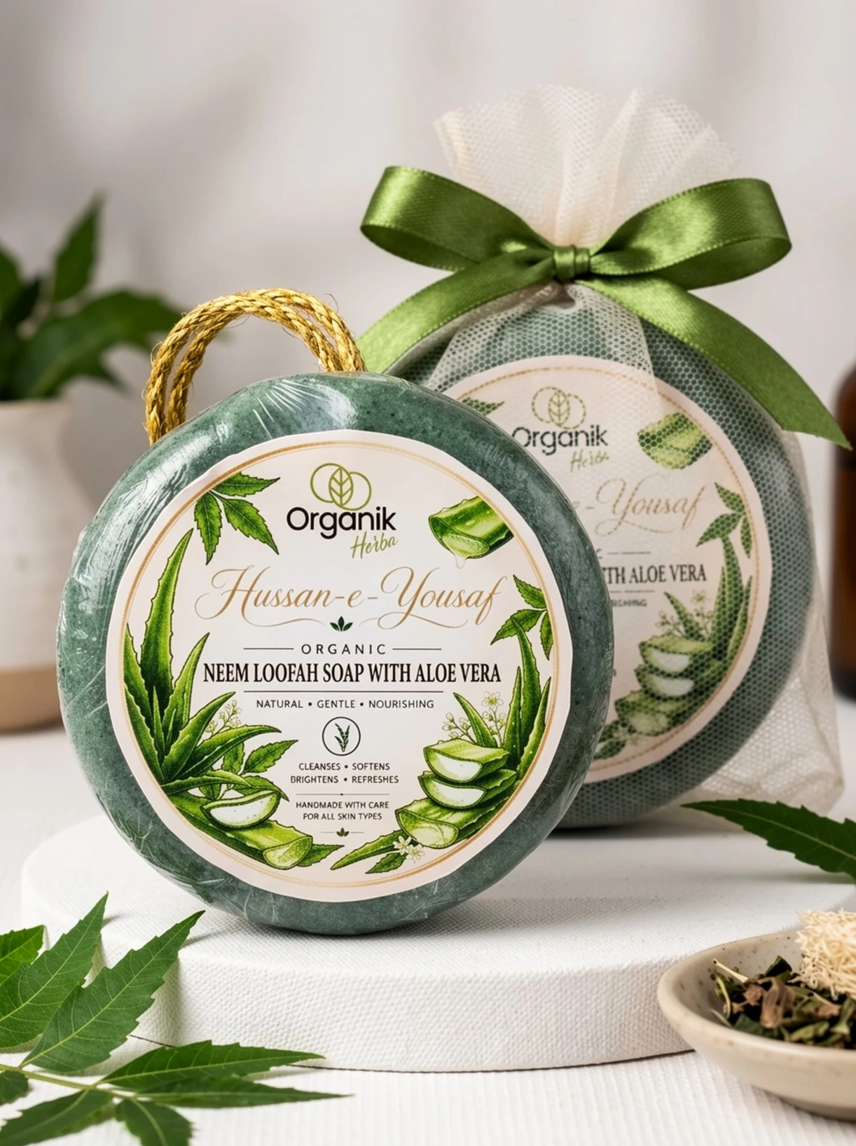 Organic handmade Neem Loofah soap with Aloe Vera by Organik Herba. The green artisanal soap bar features a botanical label with aloe illustrations, accompanied by a mesh-wrapped version tied with a green ribbon, styled with fresh neem leaves on a white textured surface.